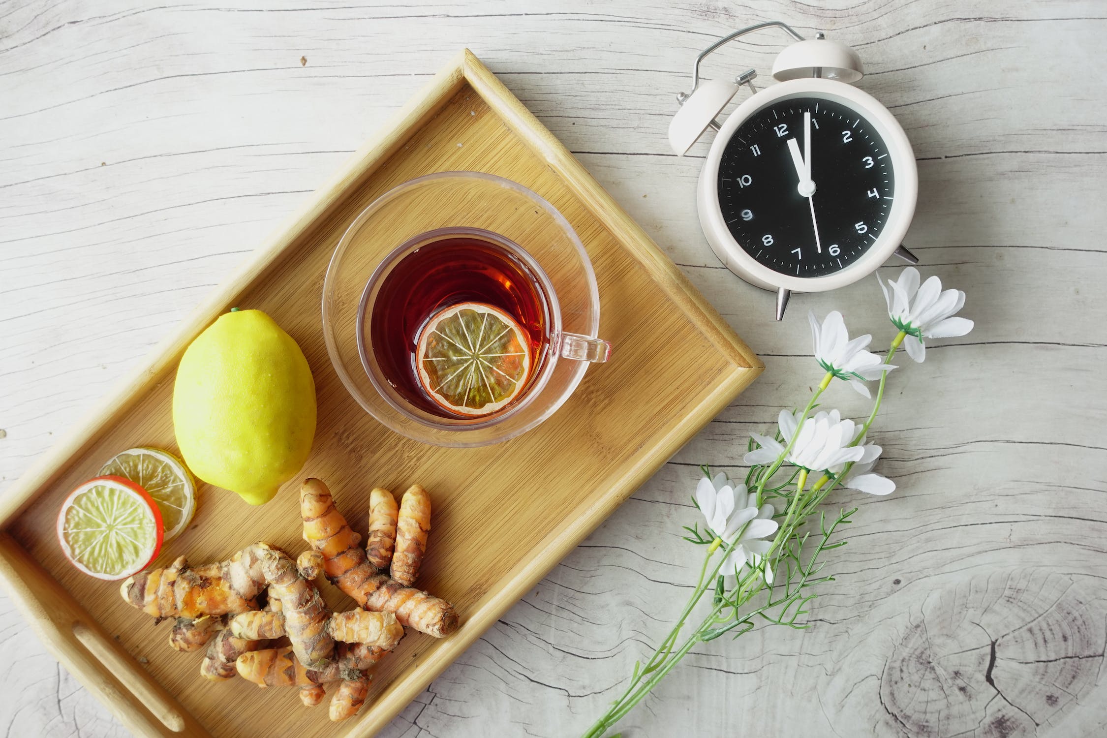 Fresh ginger & lemons on a tray with a cup of tea, next to the tray is an alarm clock and flowers